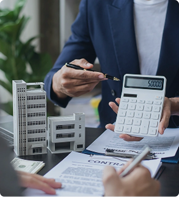 Ein Mann zeigt auf eine Zahl auf einem Taschenrechner, den er in der Hand hält. Vor ihm liegen Unterlagen und das Modell eines Hochhauses
